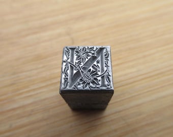 May include: A vintage metal letterpress printing block featuring the letter "K" with an ornate floral design. The block is a square shape and has a silver finish. The floral design includes detailed leaves and flowers.