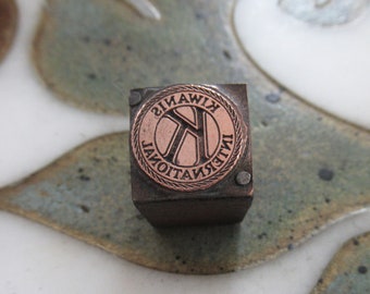 Könnte beinhalten: Ein vintage Druckblock aus Metall mit einem kupferfarbenen kreisförmigen Design. Das Design zeigt einen stilisierten Buchstaben "K" in einem Kreis mit dem Text "KIWANIS INTERNATIONAL" außen herum.