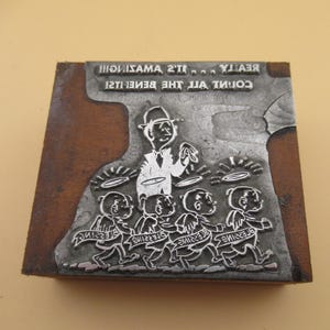 May include: A rectangular wooden printing block with a metal plate featuring an illustration of a person in a hat standing behind four small figures with wings and halos. The text reads: REALLY... IT'S AMAZING!!! COUNT ALL THE BLESSINGS!