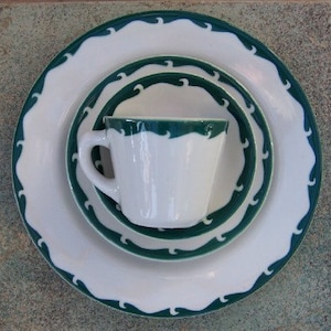 May include: A white ceramic dinner set with a dark green wave pattern. The set includes a large plate, a smaller plate, a saucer, and a teacup. The plates have a scalloped edge. The teacup has a handle.