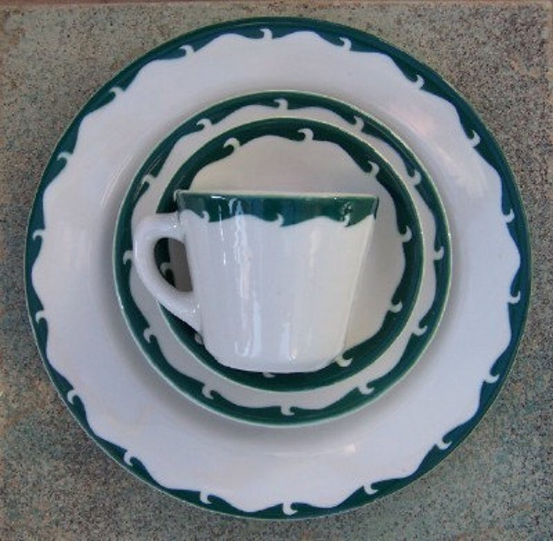 May include: A white ceramic dinner set with a dark green wave pattern. The set includes a large plate, a smaller plate, a saucer, and a teacup. The plates have a scalloped edge. The teacup has a handle.