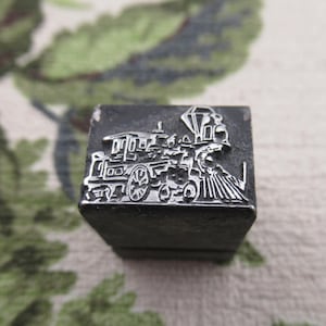 May include: A vintage metal printing block featuring a detailed relief of a steam train. The train design includes a locomotive, passenger cars, and a diamond-shaped element. The block is dark gray and sits on a patterned surface.
