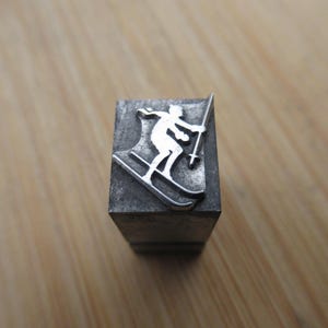 May include: A metal letterpress block featuring a skier in a downhill position. The skier is depicted in a white silhouette, holding ski poles, against a dark gray background. The block is rectangular and sits on a wooden surface.