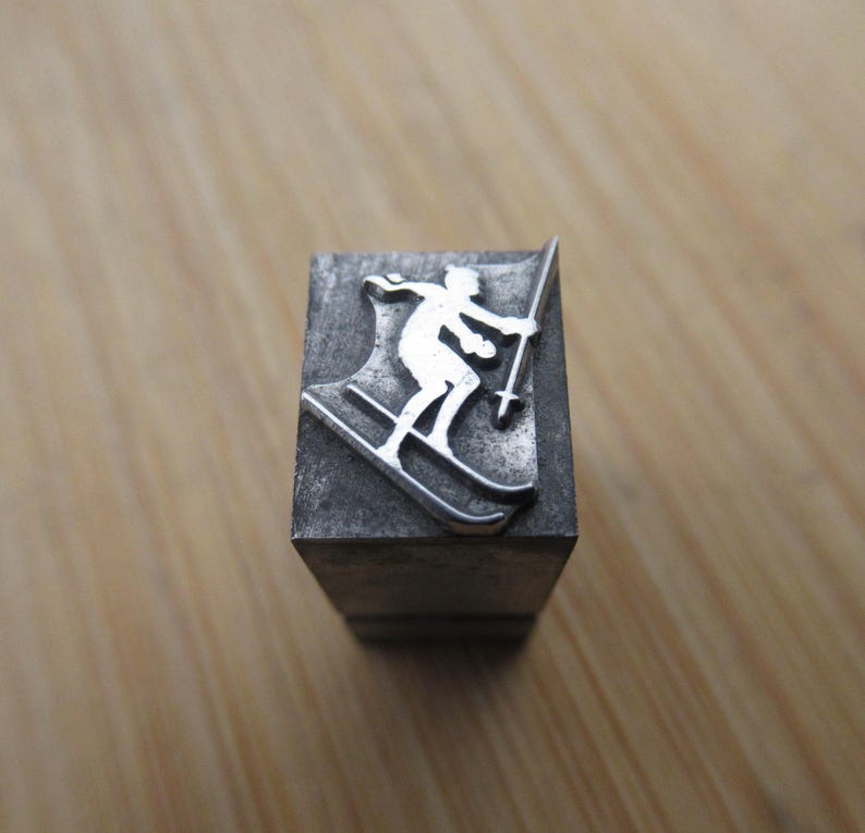 May include: A metal letterpress block featuring a skier in a downhill position. The skier is depicted in a white silhouette, holding ski poles, against a dark gray background. The block is rectangular and sits on a wooden surface.