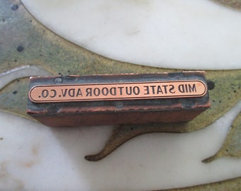 May include: A vintage metal printing block with a copper-coloured label that reads "MID STATE OUTDOOR ADV. CO."