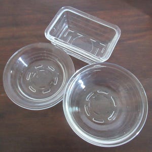 Könnte beinhalten: Ein Set aus drei klaren Glasbehältern, bestehend aus zwei runden Schalen und einer rechteckigen Schale. Jedes Stück hat eine glatte, transparente Oberfläche und ein geformtes Bodendesign mit einem kreisförmigen Muster am Glasboden.