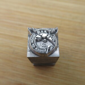 May include: A silver metal stamp featuring a moose head design with the text "ORDER OF THE WOODS" around the edge.