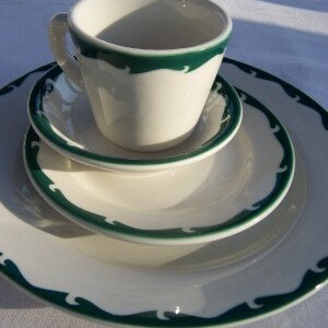 May include: A white ceramic teacup and saucer set with a green wave pattern border. The set includes a teacup, a small saucer, a medium plate, and a large plate. The set is on a white surface.