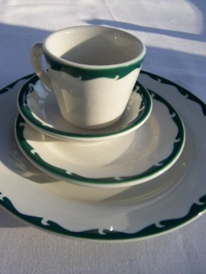 May include: A white ceramic teacup and saucer set with a green wave pattern border. The set includes a teacup, a small saucer, a medium plate, and a large plate. The set is on a white surface.