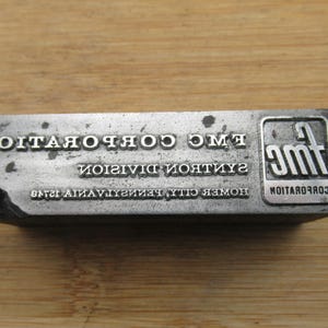May include: A rectangular metal printing block with the text "FMC CORPORATION", "SANITATION DIVISION", and "HOMER CITY PENNSYLVANIA 15748" in raised letters. The block also features the FMC logo. The block is set against a wooden surface.