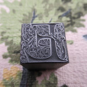 May include: Close-up of a vintage metal letterpress printing block. The letter "H" is intricately designed with floral and scrollwork details. The block is a dark gray color and appears to be made of metal, showcasing a classic, antique aesthetic.