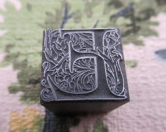 May include: Close-up of a vintage metal letterpress printing block. The letter "H" is intricately designed with floral and scrollwork details. The block is a dark gray color and appears to be made of metal, showcasing a classic, antique aesthetic.