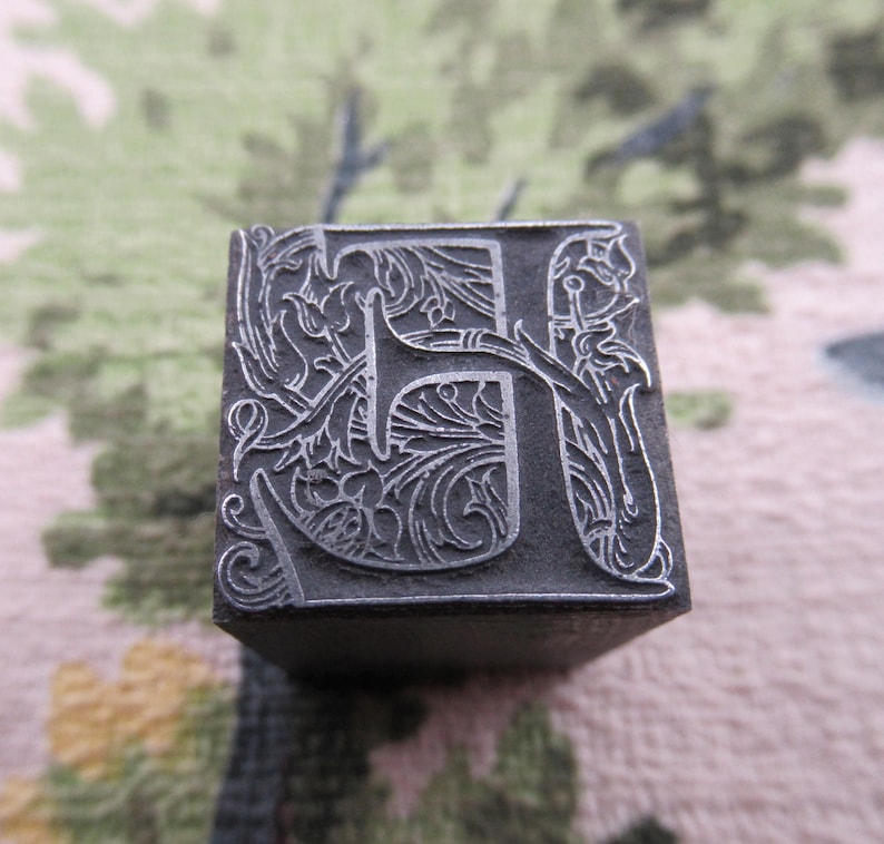 May include: Close-up of a vintage metal letterpress printing block. The letter "H" is intricately designed with floral and scrollwork details. The block is a dark gray color and appears to be made of metal, showcasing a classic, antique aesthetic.