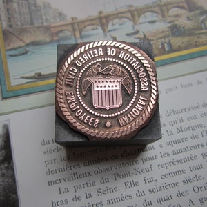 Op de afbeelding: Een koperkleurige metalen stempel met een cirkelvormig ontwerp. De stempel heeft een schild met sterren en de tekst "CIVIL EMPLOYEES NATIONAL UNION OF NOTARIES 1922".