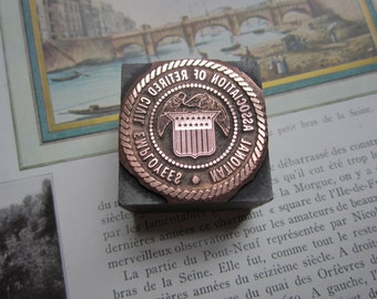 Könnte beinhalten: Ein kupferfarbener Metallstempel mit einem runden Design. Der Stempel zeigt ein Schild mit Sternen und den Text "CIVIL EMPLOYEES NATIONAL UNION OF NOTARIES 1922".