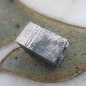 May include: A metal letterpress printing block with the letter "S" engraved on the face. The block is gray and has a worn, textured surface.