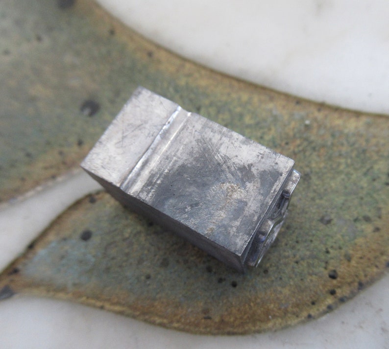 May include: A metal letterpress printing block with the letter "S" engraved on the face. The block is gray and has a worn, textured surface.