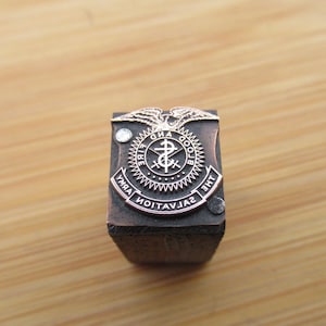 May include: A vintage Salvation Army emblem printing block. The rectangular block is made of dark wood with a detailed metal emblem on top. The emblem features an eagle, text, and a cross symbol.