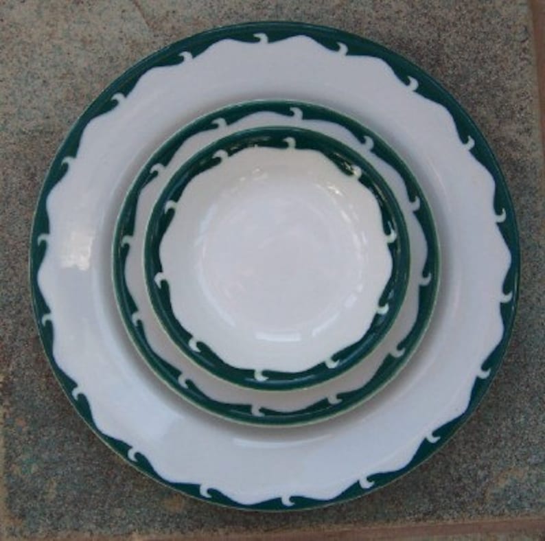 May include: A set of three white ceramic dishes with a dark green wave pattern border. The set includes a large plate, a smaller plate, and a bowl. The dishes are arranged in a nested configuration.