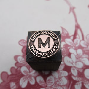 Puede incluir: Un sello de metal negro con un diseño circular. El diseño presenta el texto "MONTGOMERY POWER COMPANY" que rodea una gran letra "M".