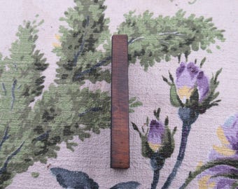 Puede incluir: Un objeto rectangular de madera marrón oscuro. La veta de la madera es visible. El objeto está colocado verticalmente sobre un fondo floral con árboles verdes y flores moradas.