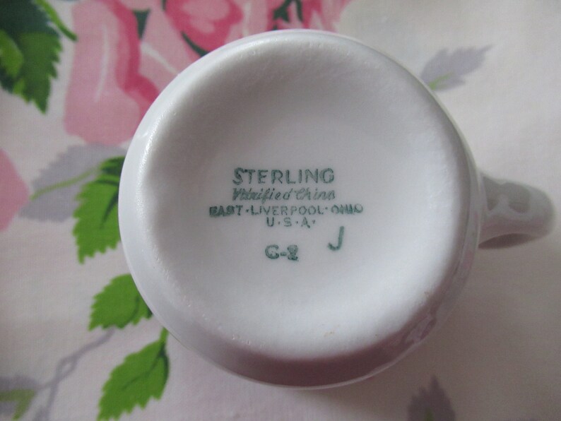 May include: A white ceramic mug, seen from below, with a handle. The base is marked with green text: "STERLING Vitrified China EAST - Liverpool, OHIO U.S.A." The mug's design is simple and classic, suitable for everyday use.