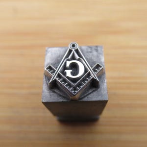 May include: A square metal printing block featuring a raised silver-toned Masonic square and compass emblem with a central letter G. The emblem is centered on the flat top surface of the block, which has a metallic gray finish.
