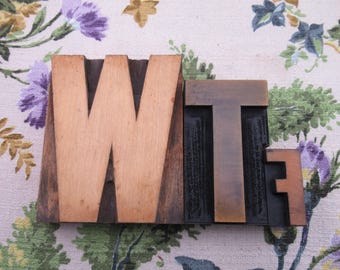 May include: Wooden letterpress printing blocks spelling out the letters "WTF". The "W" is a light brown color, while the "T" and "F" are a darker brown. The blocks are on a floral patterned fabric.