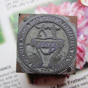 May include: A vintage metal printing block with the text "INDEPENDENT DANCE STUDIOS INC AMERICA" encircling a central design of two figures dancing. The word "AAIDA" is also visible. The block is square and has a wooden base.