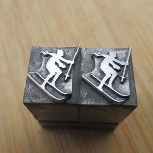 May include: Two metal printing blocks with a skier silhouette. The skier is depicted in white, holding ski poles, and positioned on skis. The blocks are rectangular and made of a dark, textured metal, likely used for printing or crafting.