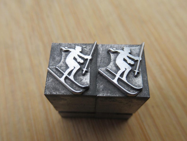 May include: Two metal printing blocks with a skier silhouette. The skier is depicted in white, holding ski poles, and positioned on skis. The blocks are rectangular and made of a dark, textured metal, likely used for printing or crafting.