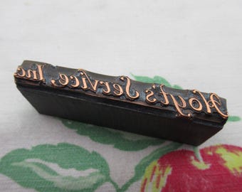 May include: A vintage letterpress stamp with the text "Hodge's Service, Inc." in copper-coloured raised lettering. The stamp is made of dark wood and is angled slightly, revealing the text. The background is a floral pattern.