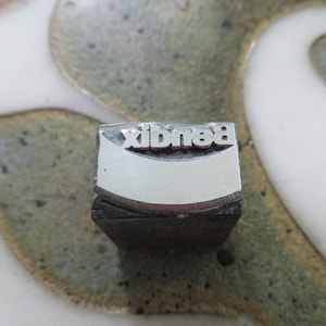 May include: A metal printing block with the word "Bendi" embossed on it. The block is grey and has a slightly curved surface.