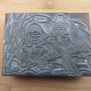 May include: A vintage metal printing block featuring a relief design of two figures, one wearing a hat, with a stylised background of leaves and branches.
