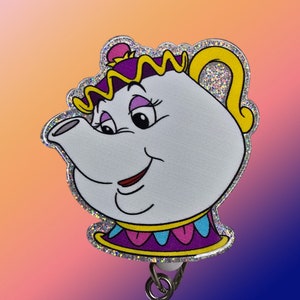 Pode incluir: Um personagem de chaleira sorridente com a forma de uma chaleira, com glitter prateado, roxo e amarelo. O personagem é um personagem popular da Disney do filme A Bela e a Fera.