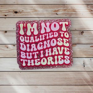 Op de afbeelding: Roze glitter sticker met de tekst "Ik ben niet gekwalificeerd om te diagnosticeren, maar ik heb theorieën" in een retro-lettertype.