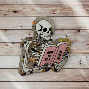 May include: A skeleton reading a book titled "How to be a Nurse". The book is pink with gold lettering. The skeleton is white with black outlines and is sitting on a glittery background with black and gold glitter.
