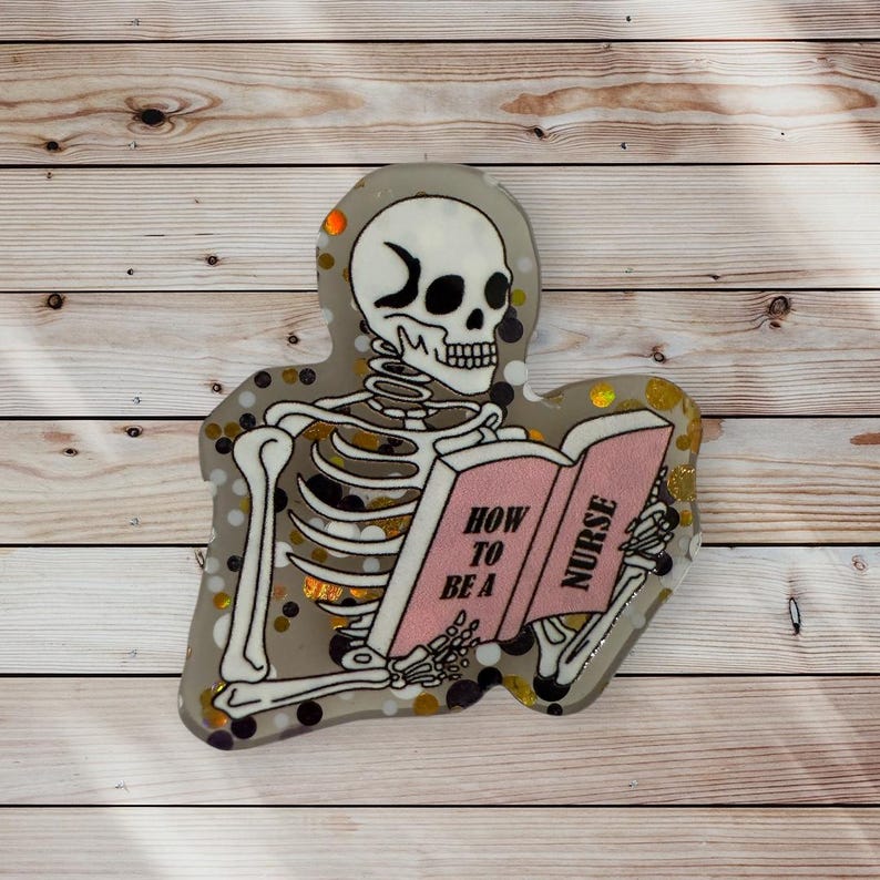 May include: A skeleton reading a book titled "How to be a Nurse". The book is pink with gold lettering. The skeleton is white with black outlines and is sitting on a glittery background with black and gold glitter.