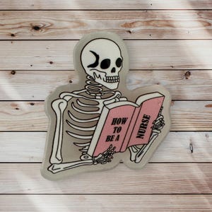 May include: A white skeleton with a crescent moon on its forehead is reading a pink book titled "How to be a Nurse".