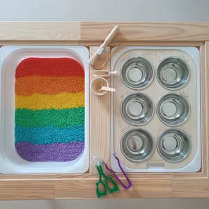 Puede incluir: Una mesa de juego de madera con dos compartimentos. Un compartimento tiene una bandeja blanca llena de arroz de colores arcoíris. El otro compartimento tiene una bandeja blanca con seis cuencos de acero inoxidable etiquetados como "Grün", "Lila", "Blau", "Rot", "Orange" y "Lena & Tim".