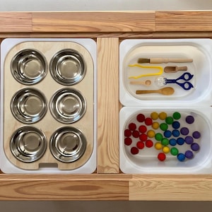 Op de afbeelding: Een houten speeltafel met drie compartimenten. Het linker compartiment heeft negen kleine metalen kommen. Het middelste compartiment heeft een houten lepel, een houten borstel, een gele pincet, een klein glazen flesje en een blauwe schaar. Het rechter compartiment heeft kleurrijke ballen.