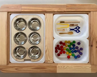 Könnte beinhalten: Ein hölzerner Spieltisch mit drei Fächern. Das linke Fach hat neun kleine Metallschalen. Das mittlere Fach hat einen Holzlöffel, eine Holzbürste, eine gelbe Pinzette, eine kleine Glasflasche und eine blaue Schere. Das rechte Fach hat bunte Bälle.