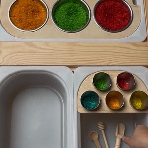 Può includere: Un set da gioco sensoriale con riso colorato in ciotole di metallo, disposto su un vassoio di legno. Il set include piccole palette e cucchiai di legno. I colori del riso includono arancione, verde, rosso e giallo.