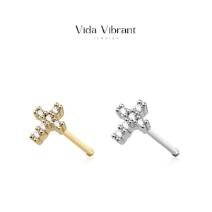 May include: Two nose rings with a cross design set with clear cubic zirconia stones. The nose ring on the left is gold and the nose ring on the right is silver.