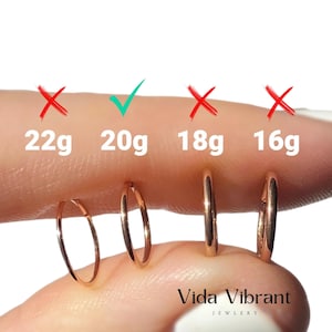 May include: Close-up of a finger wearing four different sizes of gold hoop earrings. The earrings are labeled with their gauge sizes: 22g, 20g, 18g, and 16g. A red X marks the 22g and 18g earrings, while a green check mark indicates the 20g earring is the correct size. The text "Vida Vibrant Jewelry" is visible at the bottom of the image.