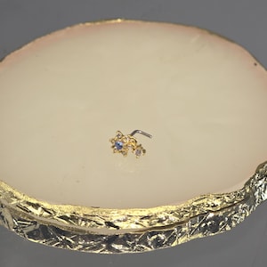 May include: A gold-coloured nose ring with a cluster of small, sparkling gemstones. The design includes a central flower-shaped element with a blue gemstone, and smaller, clear gems. The nose ring is set against a light-coloured background.