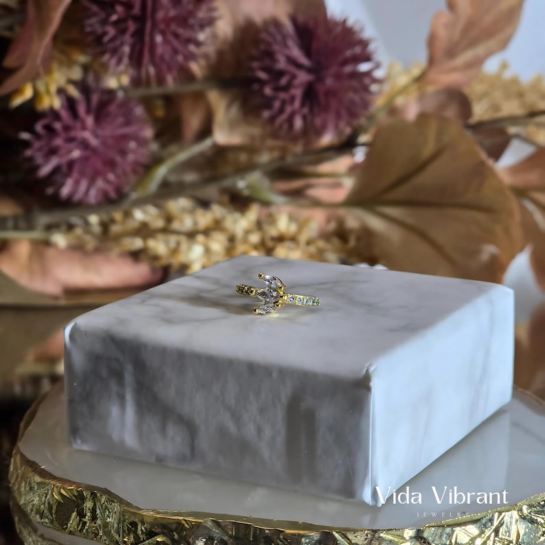May include: A gold ring with a crescent moon design and clear cubic zirconia stones. The ring is on a white marble surface.