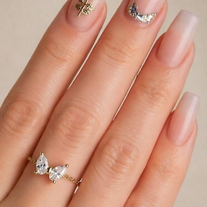 May include: A gold ring featuring two pear-shaped clear gemstones in a butterfly design. The ring is on a hand with long, light pink nails. The nails have gold and crystal nail art. The text "Vida Vibrant Jewelry" is visible.