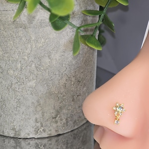 May include: A gold nose stud with a floral design, featuring small clear gemstones. The stud is shown on a person's nose, with a blurred background of a plant and a concrete pot.
