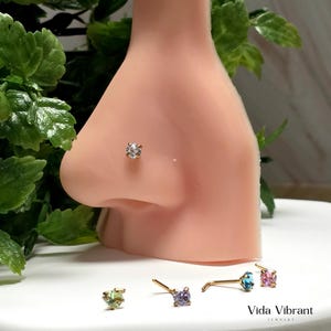May include: A gold-toned nose stud with a clear, round gemstone is shown on a mannequin nose. Several other nose studs with different coloured gemstones are arranged below. The brand name "Vida Vibrant Jewelry" is visible.